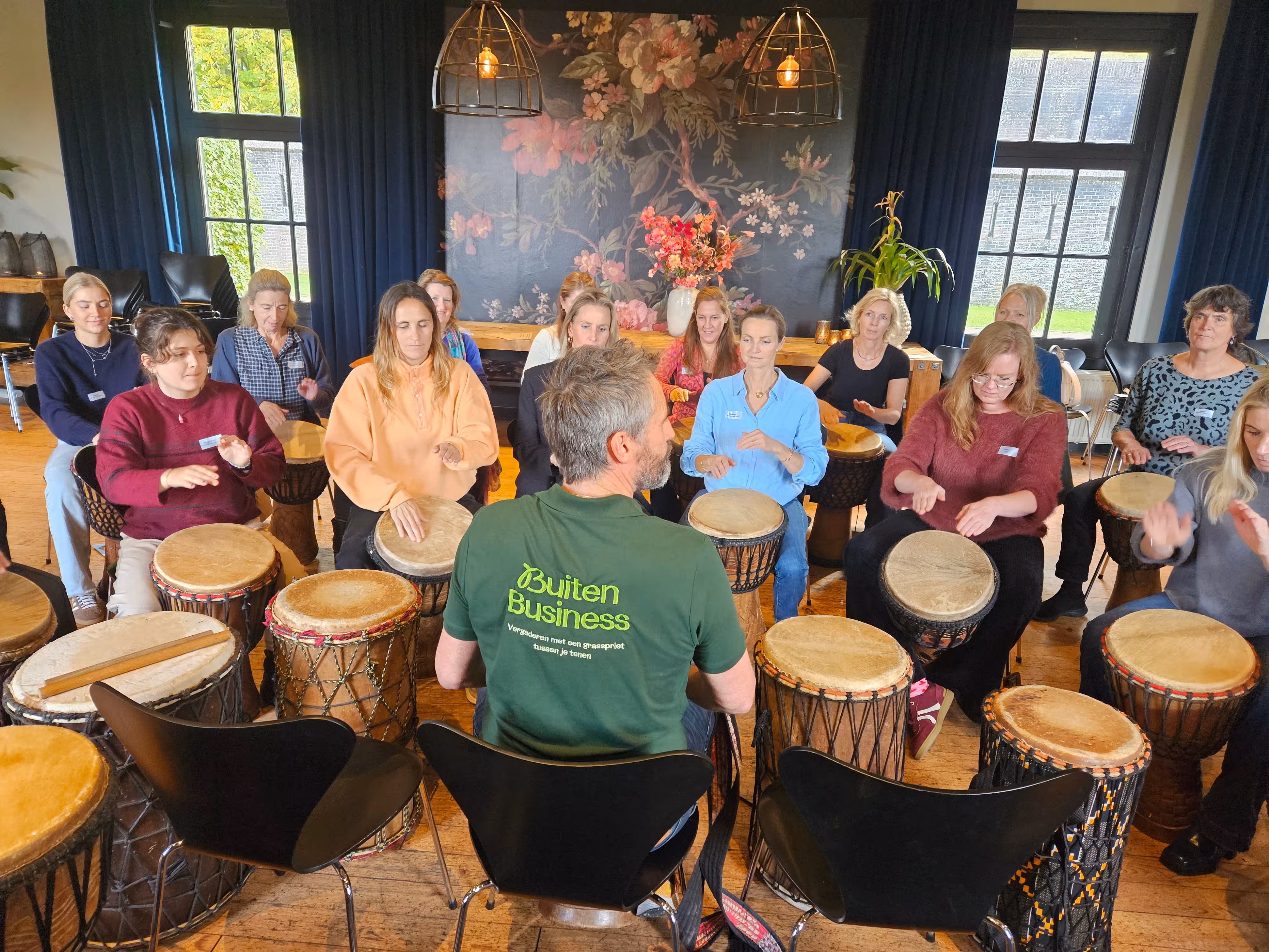 Binnenruimte met veertien personen die rondom de BuitenBusiness instructeur tijdens de Percussie Workshop