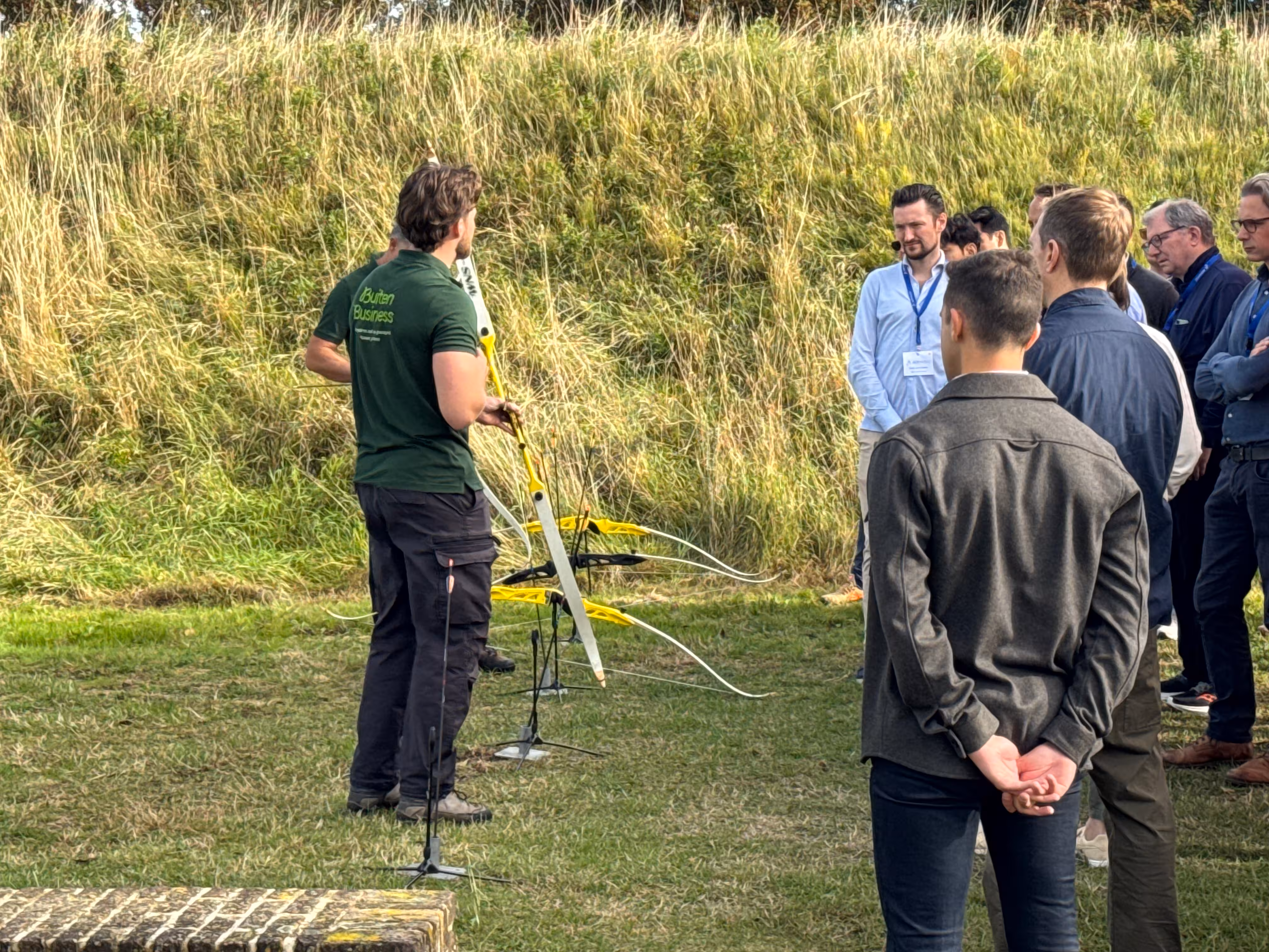 De BuitenBusiness instructeur geeft instructies en staat bij de pijl en bogen. De deelnemers luisteren aandachtig en staan op een grasveld
