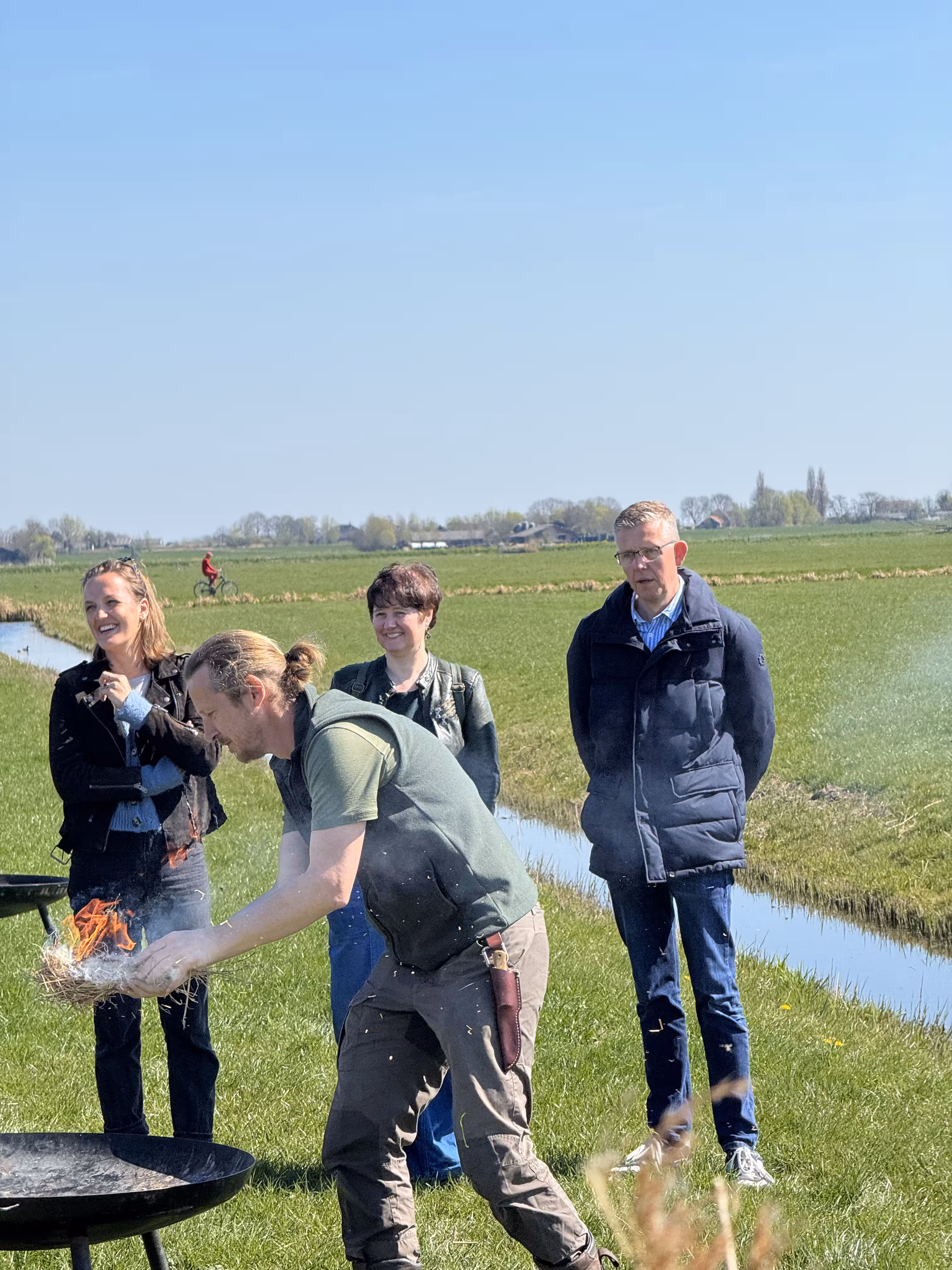 De BuitenBusiness instructeur legt een brandend vuur in de vuurschaal. De deelnemers kijken toe en staan op een grasveld met een slootje