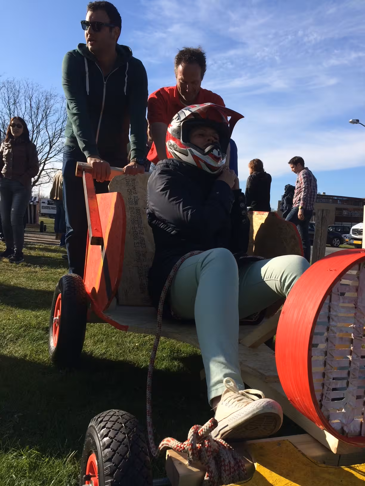 Een rode zeepkist met daarachter twee mannen die duwen. Op de rode zeepkist zit een vrouw met een zwart, wit en rode helm