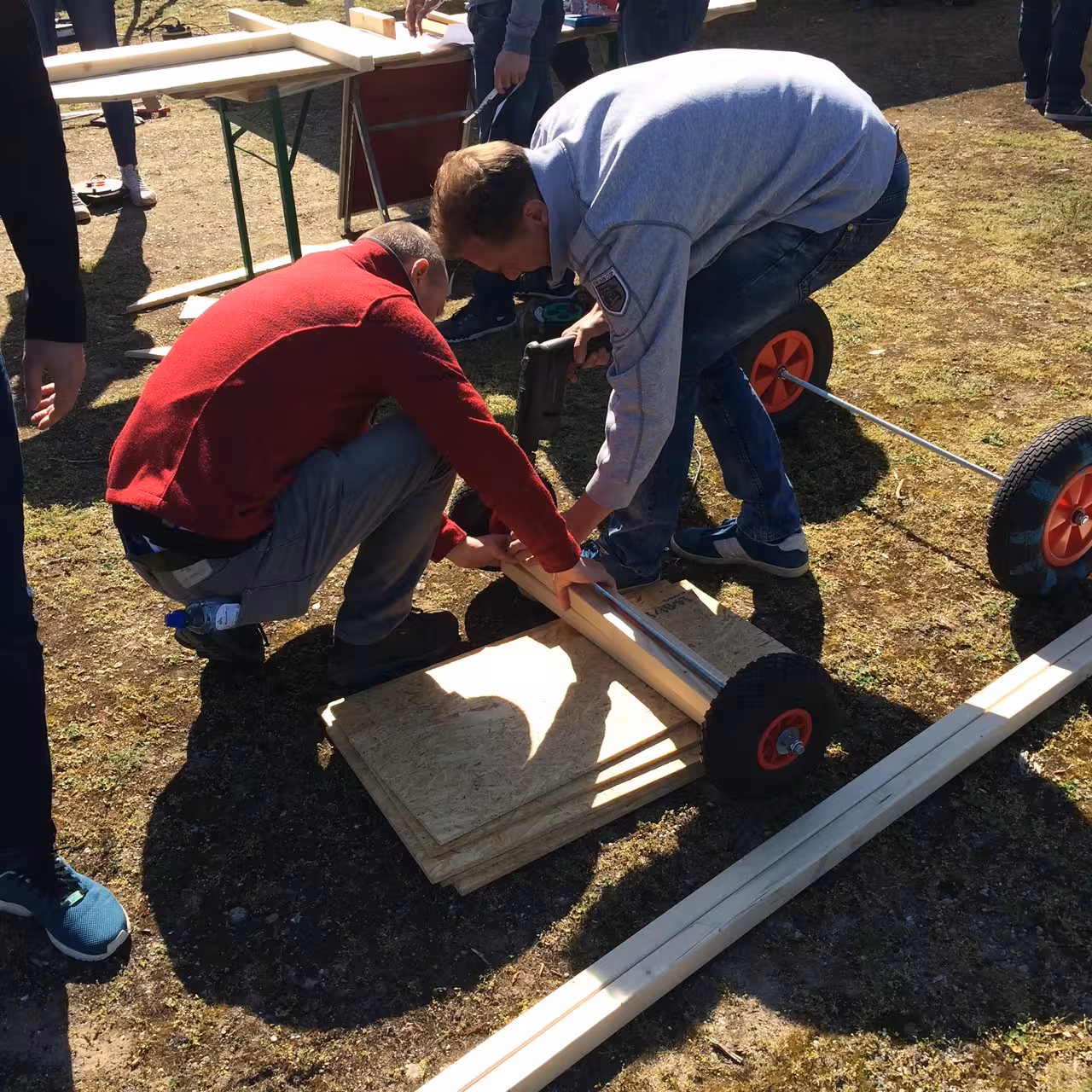 Twee mannen knielen op een grasveld bij houten platen en wielen. De man met een grijze sweater heeft een boormachine vast.