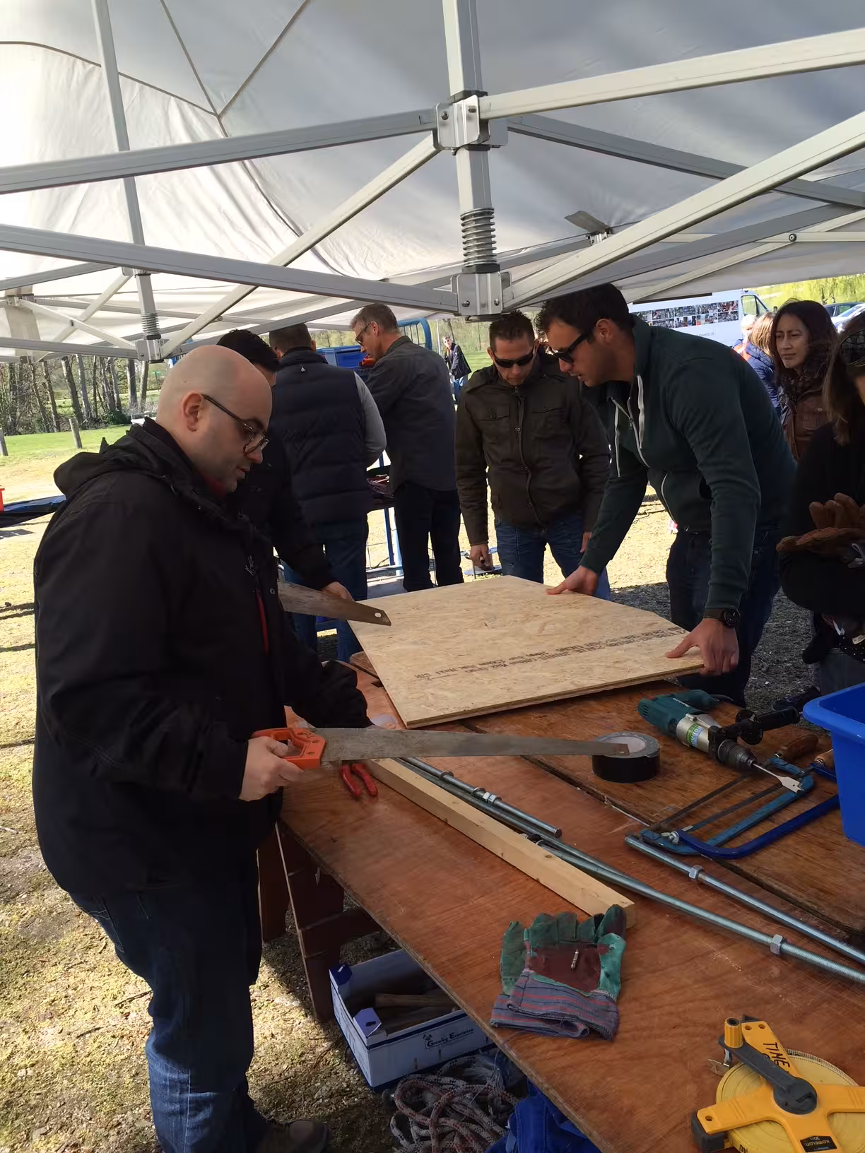 Onder een witte tent gaat een team een plaat hout zagen tijdens de zeepkisten race van buitenbusiness