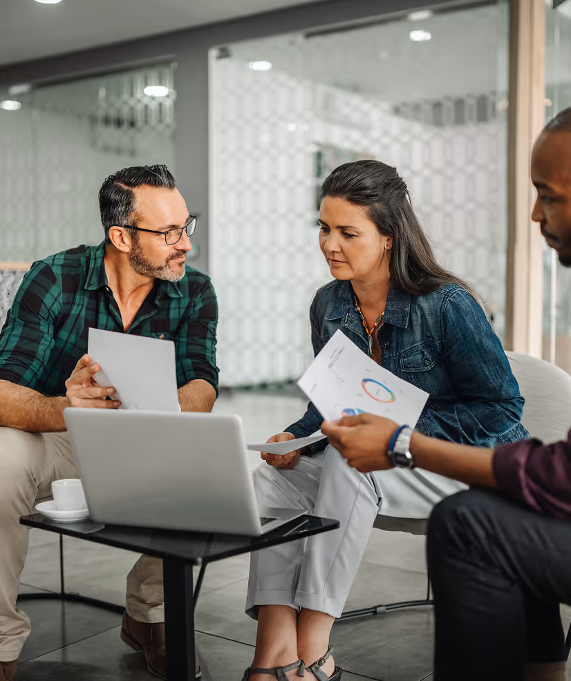 Three professionals in a modern office reviewing documents and discussing work around a laptop on a small table.