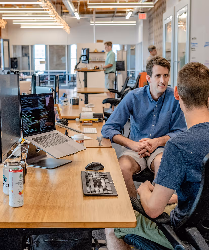 Two people sit and talk at a shared desk in a modern office with computers, while others work in the background.