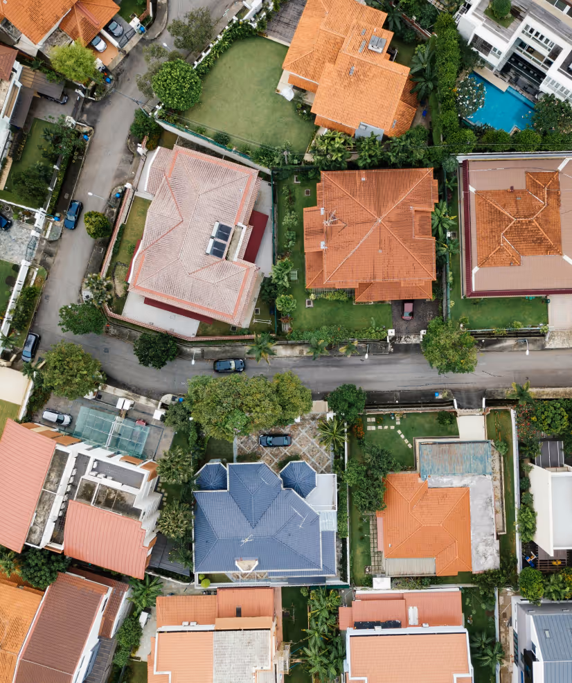 Aerial view of a suburban neighborhood with houses featuring orange and blue rooftops, green lawns, and cars parked along narrow streets.