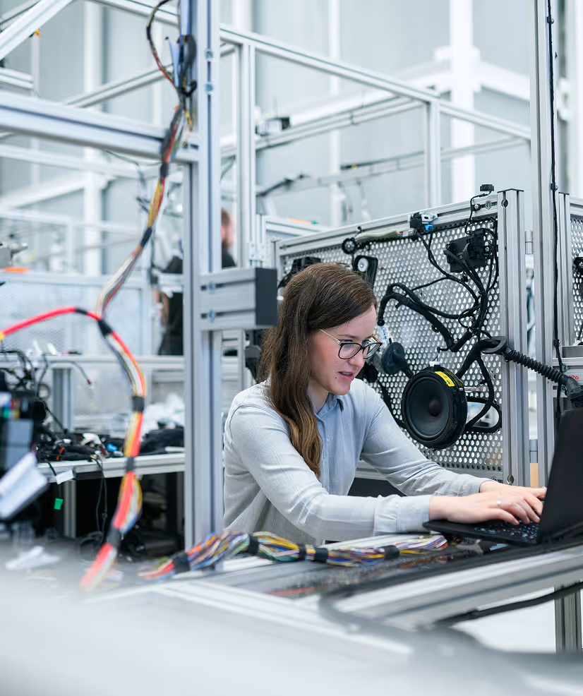 Woman wearing glasses working on a laptop in a high-tech industrial or manufacturing setting with wiring and electronic components.