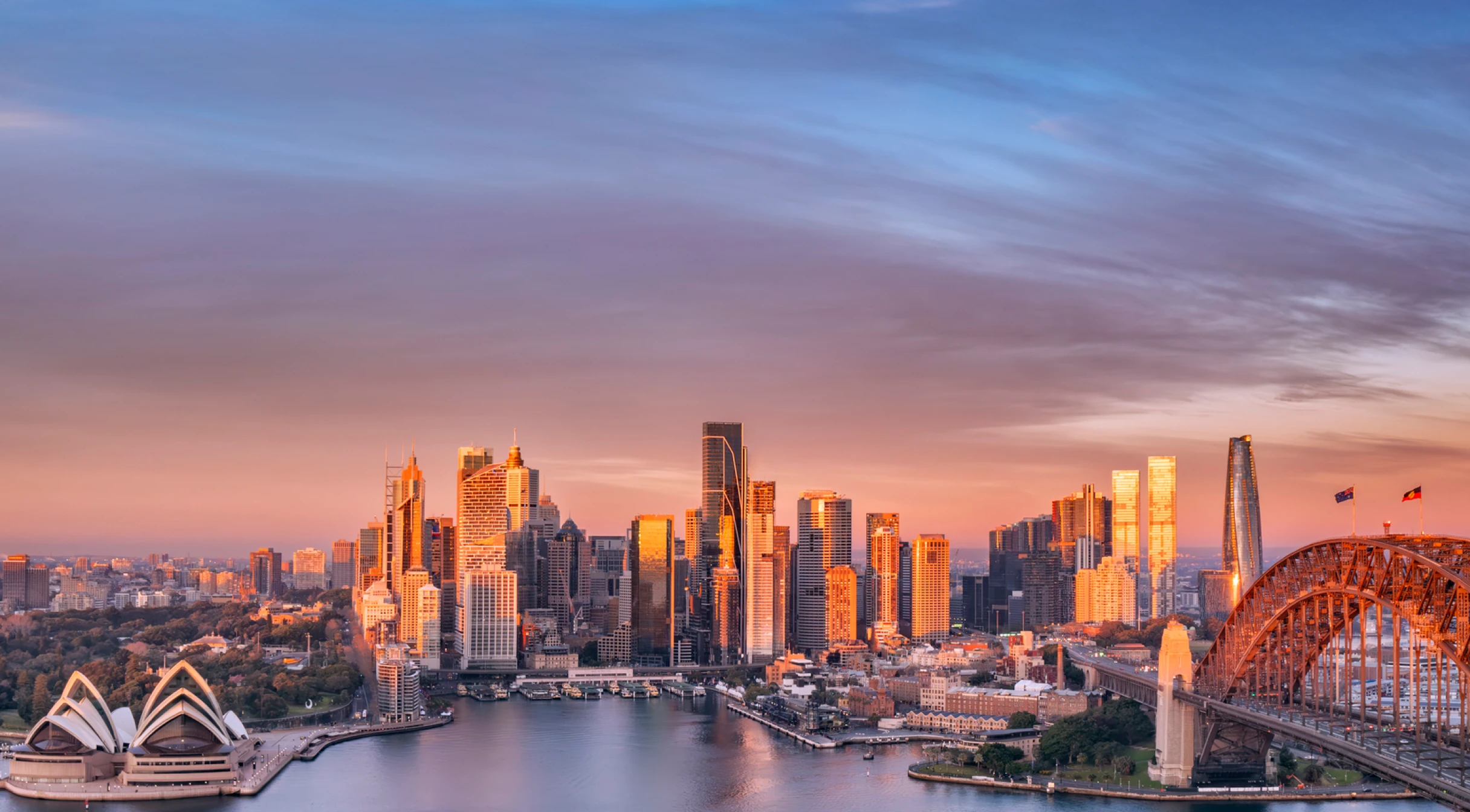 Sydney skyline at sunset with Opera House on left and Harbour Bridge on right over water.