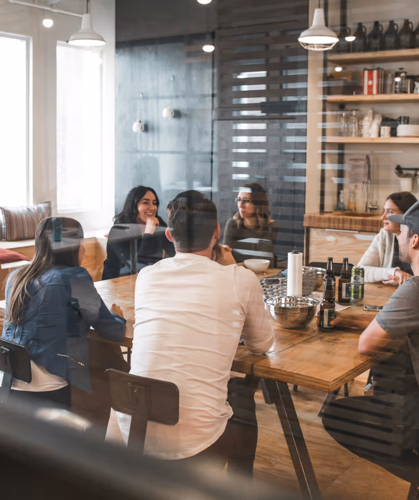 Group of six young adults sitting around a wooden table in a modern kitchen having a conversation.