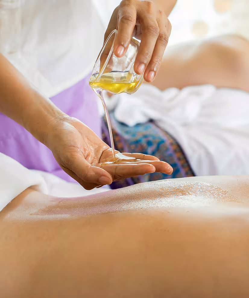 Hand pouring massage oil from a small glass container onto another hand above a person's back.
