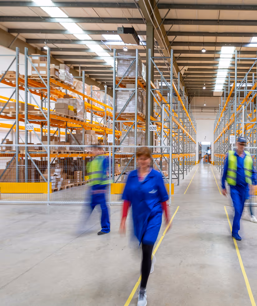 Warehouse interior with tall shelves and three workers in blue uniforms walking down the aisle.