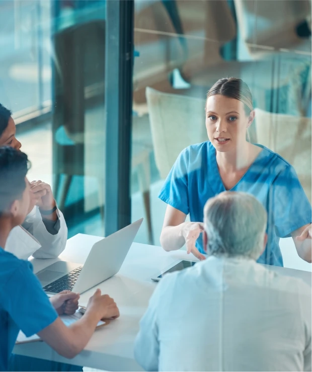 Healthcare professionals in scrubs and lab coats hold a discussion around a table with a laptop.