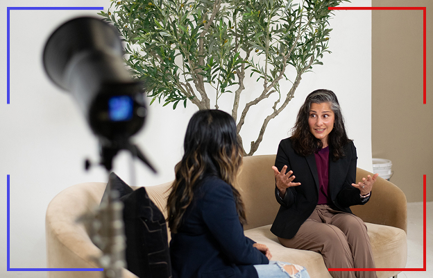 Two women sitting on a light beige sofa having a conversation with a camera set up in the foreground and a small tree behind them.