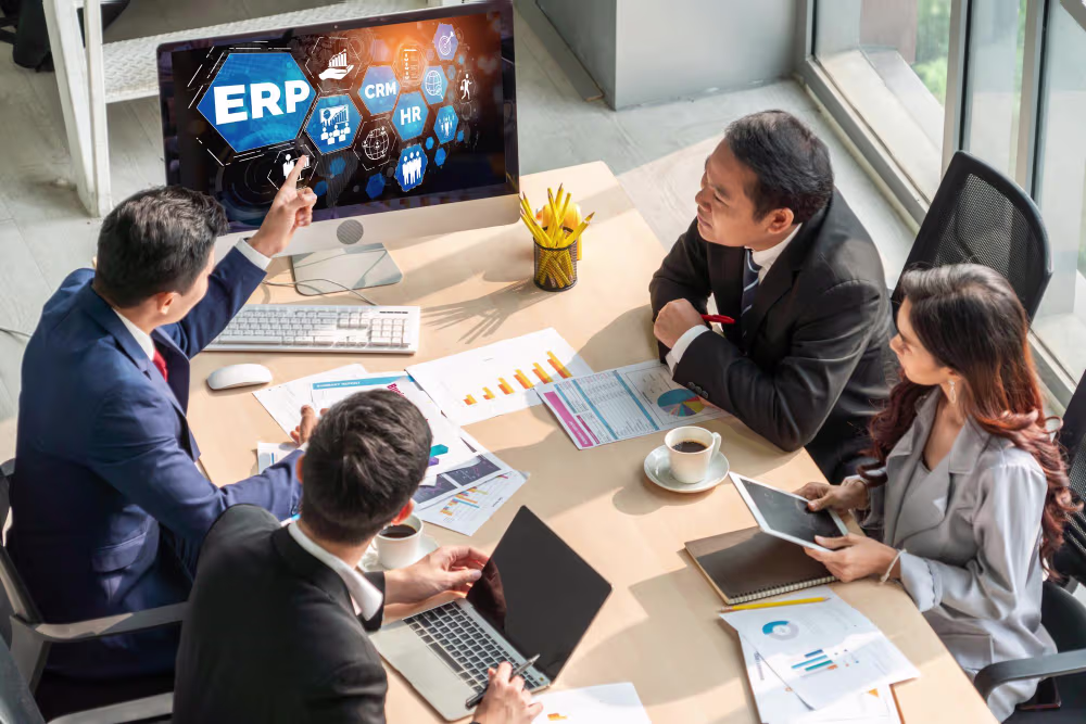 A group of four NetSuite Technical Consultants sitting together at a desk pointing to a monitor with ERP, CRM, and HR displayed on the screen.