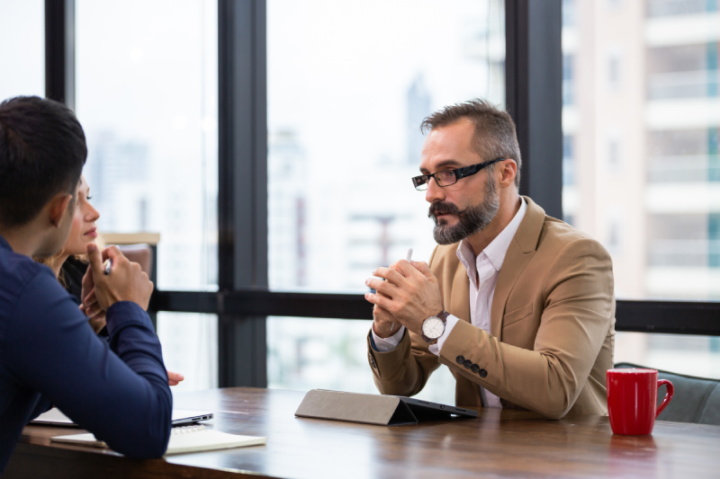 A man in a tan blazer and glasses is engaged in a discussion with two people at an office table. A red mug and tablet are on the table. The mood is focused.