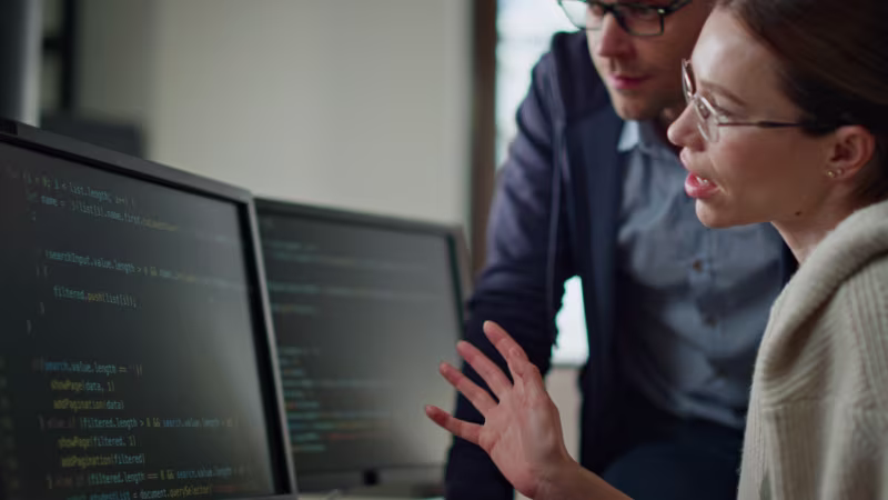 Two people, a woman and a man, are intently discussing code on a computer screen. The atmosphere is focused and collaborative.