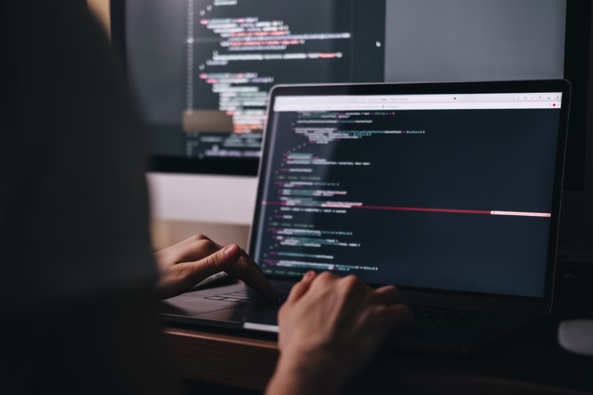 A person types code on a laptop, with colorful lines of code on the screen. A larger monitor in the background also displays coding work, creating a focused, tech-driven atmosphere.