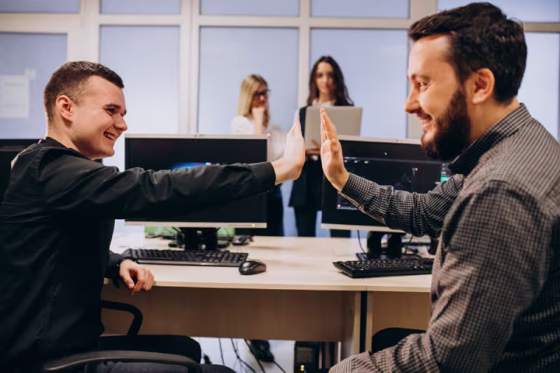 Two men in an office, sitting at desks with computers, share a high-five, smiling. Two women stand in the background, one using a laptop. Positive, collaborative atmosphere.