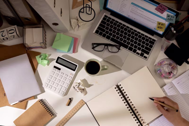 Overhead view of a cluttered desk with a laptop, open notebook, calculator, coffee, glasses, sticky notes, and papers. A hand writes in the notebook.