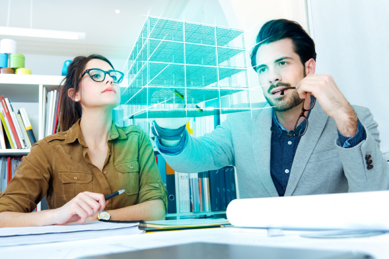 Two architects discussing a digital building model. A man in a gray blazer holds a pen, while a woman in glasses listens intently. The scene conveys collaboration and focus.