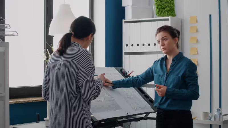 Two women are discussing architectural plans at a drafting table in a modern office. One points at documents, while the other listens intently. The atmosphere is focused.