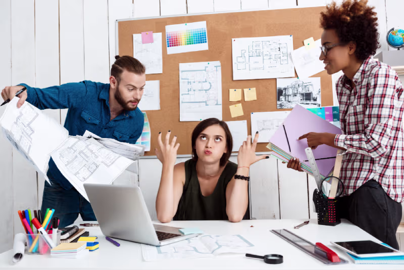 Three colleagues discuss challenges at a desk cluttered with papers and a laptop in a bright office setting.