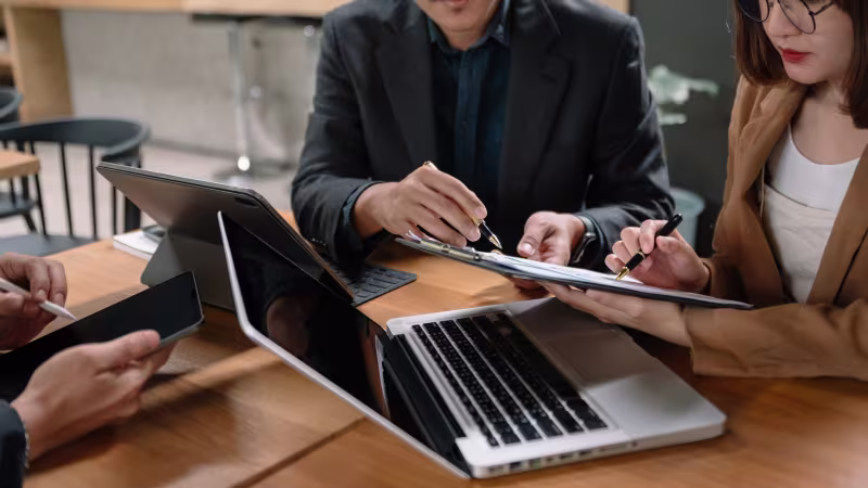 Three business professionals collaborate at a table with laptops, discussing NetSuite consultant expertise for project success.