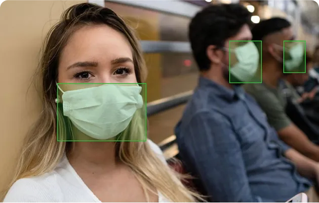 A woman in the foreground and two men in the background sitting side by side on a bench, all wearing green protective face masks.