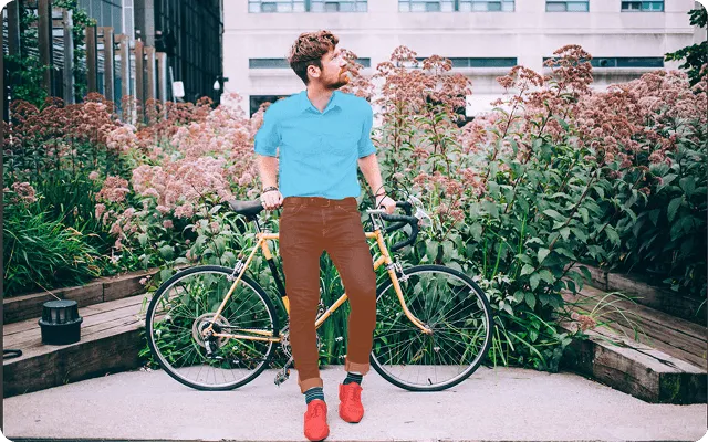 Man with a beard standing beside a yellow bicycle on a paved path surrounded by dense flowering plants and greenery.