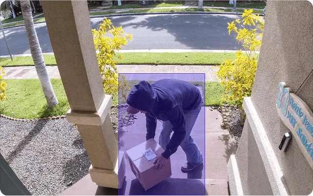 Person in a blue hoodie placing a cardboard box on a front porch near the entrance of a house.