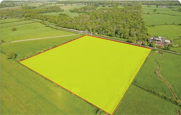 Aerial view of a large rectangular green field outlined in red, surrounded by other fields, trees, and a few houses.