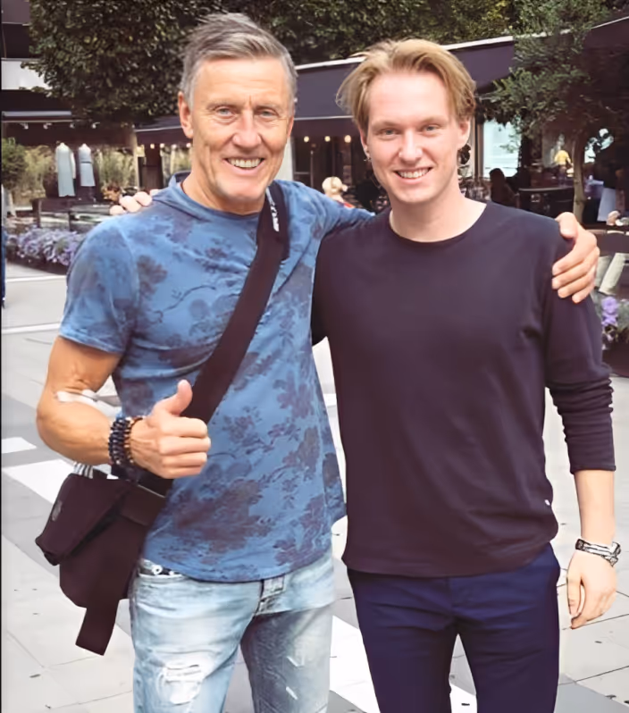 Två leende män står nära varandra utomhus, den äldre mannen visar tummen upp.