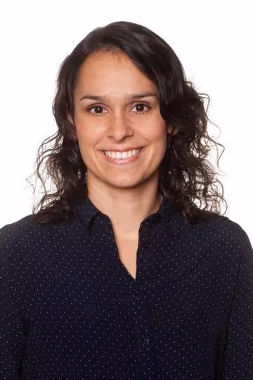 Leende kvinna med mörkt lockigt hår i svart prickig skjorta mot vit bakgrund.