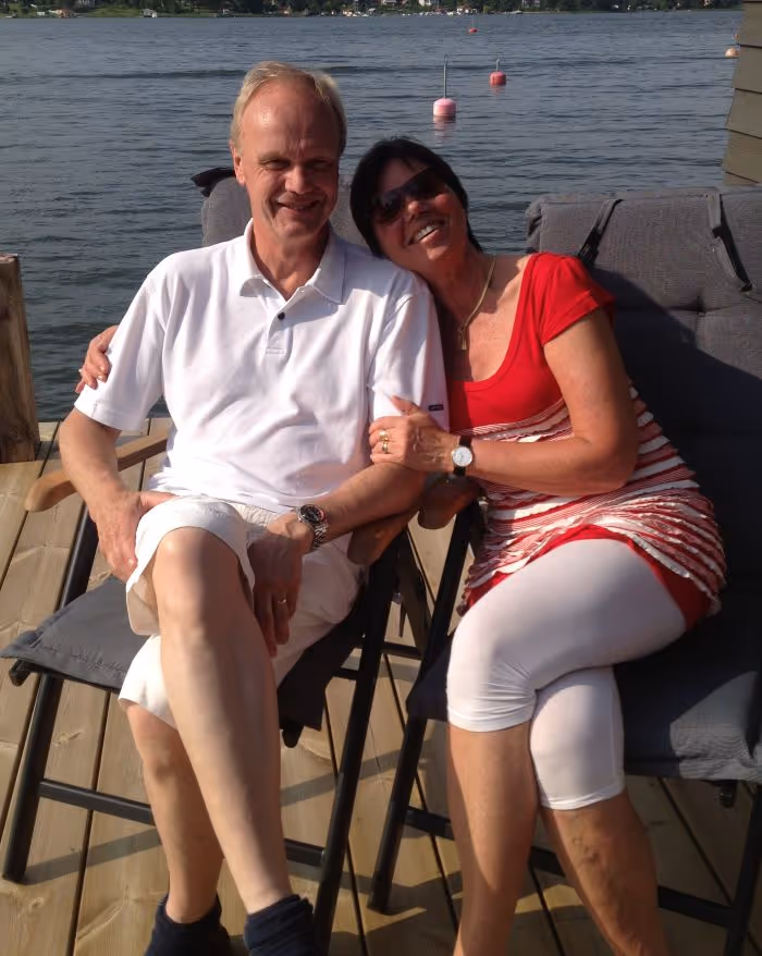 Man i vit pikétröja och kvinna i röd topp och vita leggings som sitter tillsammans och ler vid en sjö.