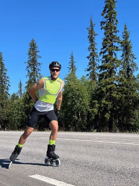 Man rollerblading on a road wearing a helmet, sunglasses, a yellow reflective vest, black shorts, and neon yellow socks with trees and blue sky in the background.