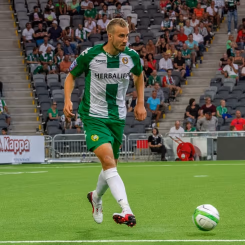 Fotbollsspelare i grönvit tröja på konstgräsplan med boll framför sig under match.