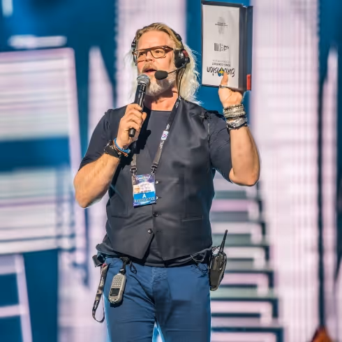 Man med långt blont hår, glasögon och headset håller mikrofon i ena handen och ett dokument i den andra på scen.