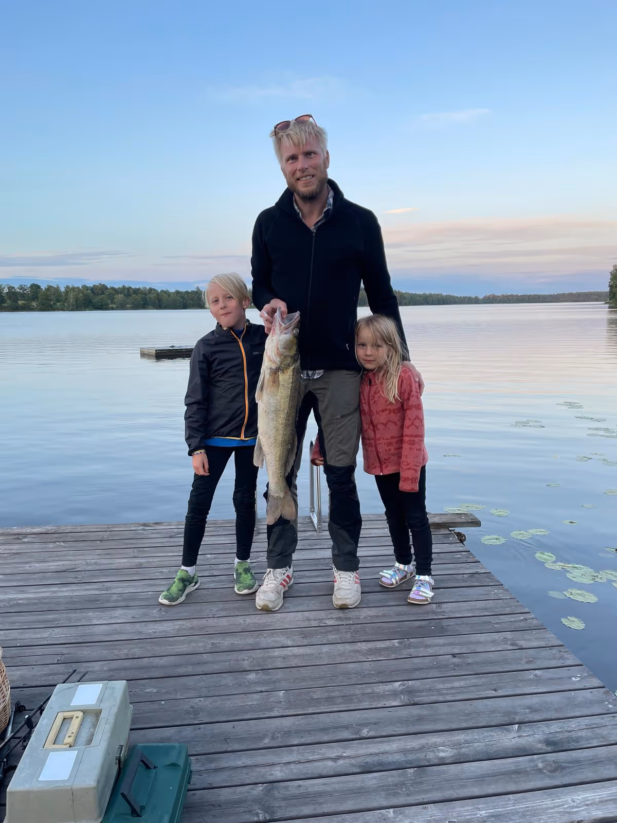 Man och två barn står på en brygga vid en sjö, mannen håller en stor fisk.