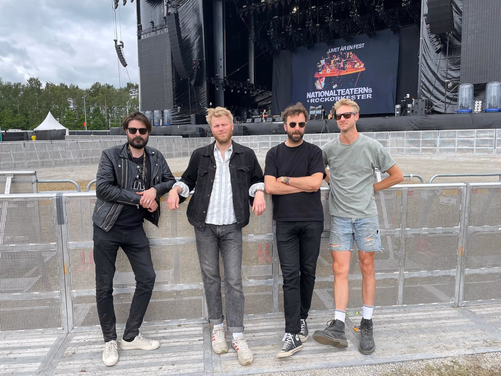 Fyra män med solglasögon lutade mot säkerhetsbarriär framför stor konsertstage med texten Nationalteaterns Rockorkester.