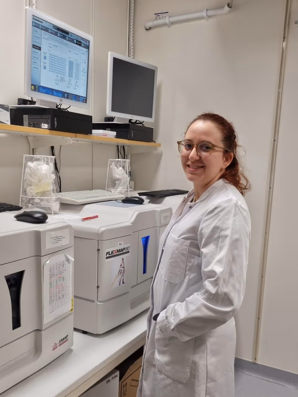 Kvinna i glasögon och vit labbrock ler bredvid avancerad laboratorieutrustning med skärmar i ett laboratorierum.