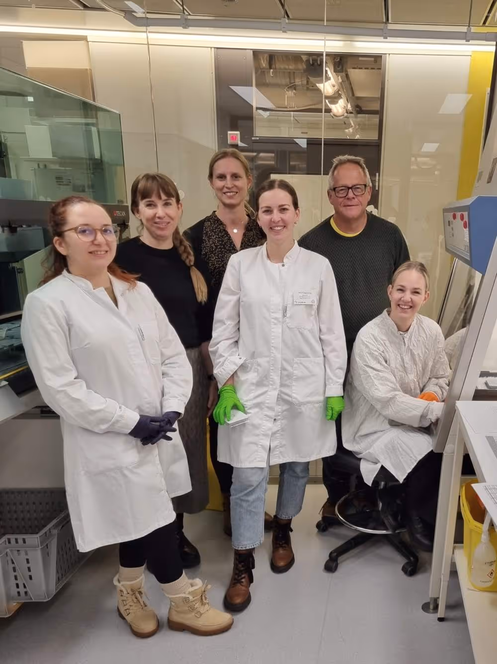 Sex personer i ett laboratorium, fyra kvinnor i labbrockar och två personer i vanlig klädsel, alla ler mot kameran.