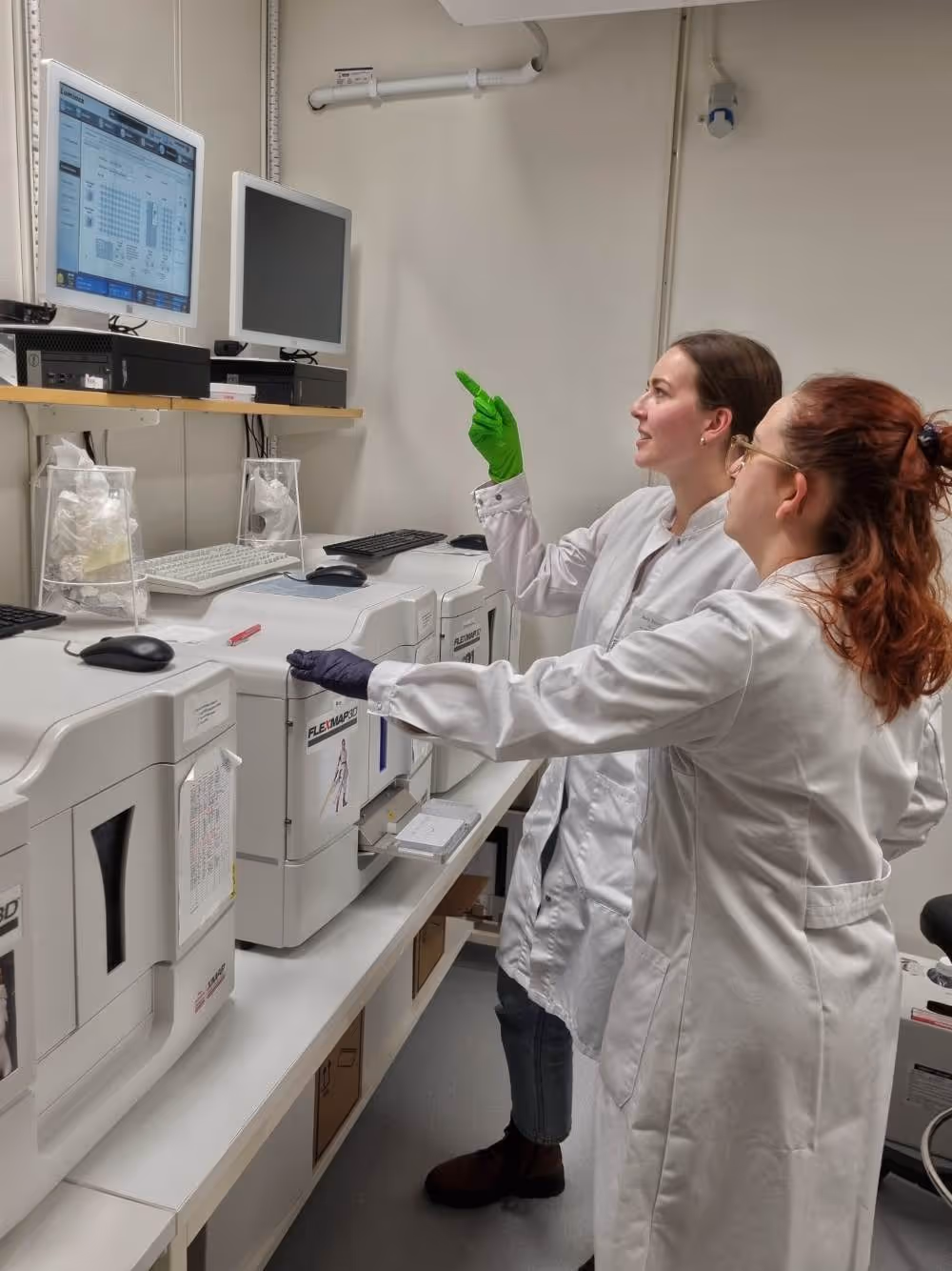 Två kvinnliga forskare i labbrockar arbetar vid och pekar på datorskärmar i ett laboratorium med avancerad laboratorieutrustning.