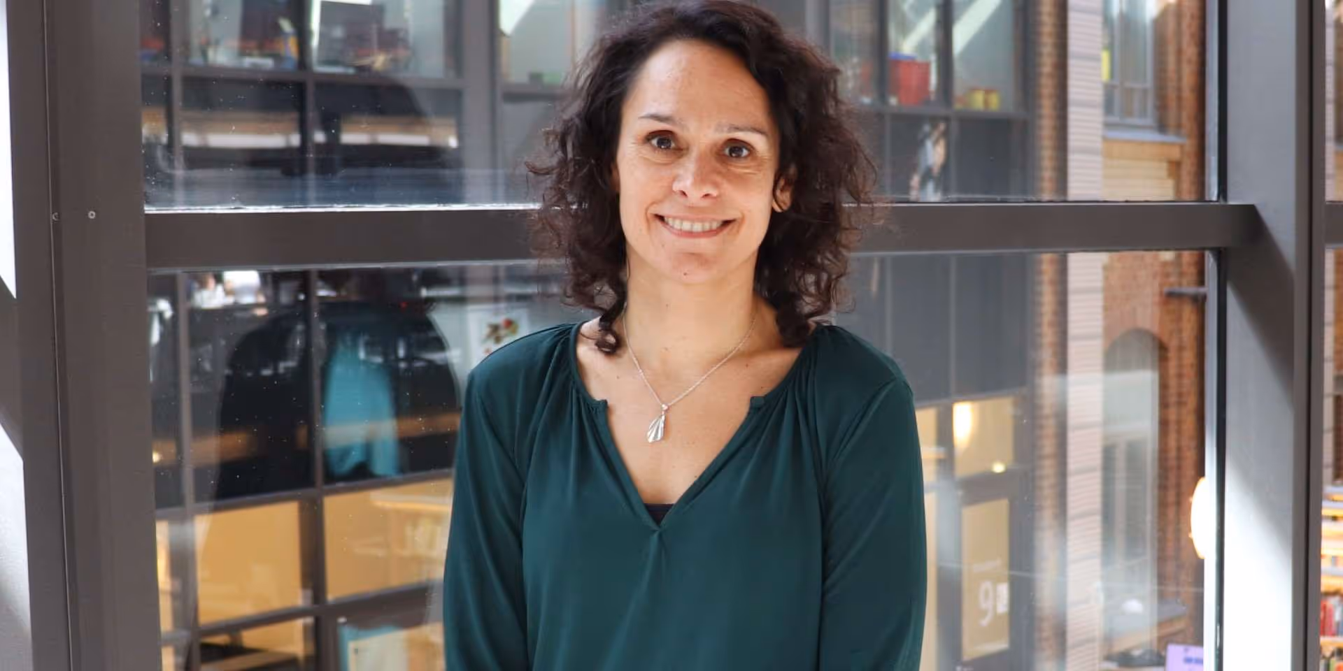 Kvinna med mörkt, lockigt hår och grön topp ler framför ett stort fönster i en modern byggnad.