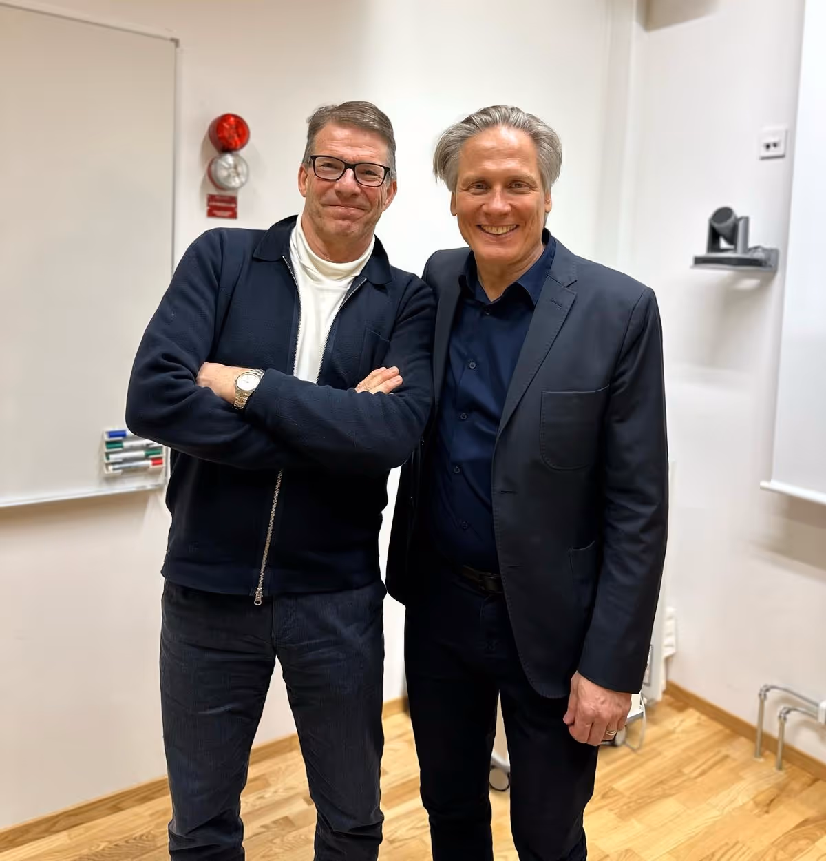 Två män står sida vid sida inomhus, den ena med armarna i kors och den andra med armen runt hans axel, båda ler mot kameran.