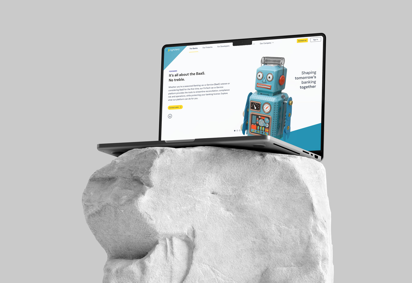 A laptop sits open with a webpage showing a blue toy robot and a tagline "it's all about the BaaS". The webpage and site promote a FinTech startup with the tagline "Shaping tomorrow's banking together"