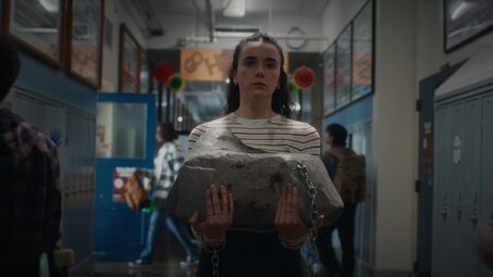 Student walks down a school hallway holding a large rock wrapped in chains, looking tense, with lockers and classmates in the background