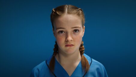 Girl with braided pigtails in a blue uniform stares ahead against a solid blue background