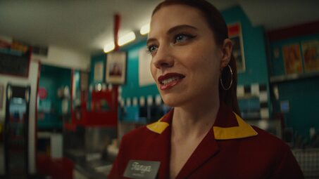 Diner server in a red-and-yellow uniform with hoop earrings speaks inside a retro-style diner, menu boards visible behind her