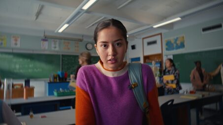 Teen in a purple-and-orange sweater looks straight at the camera in a classroom with chalkboards and desks behind her