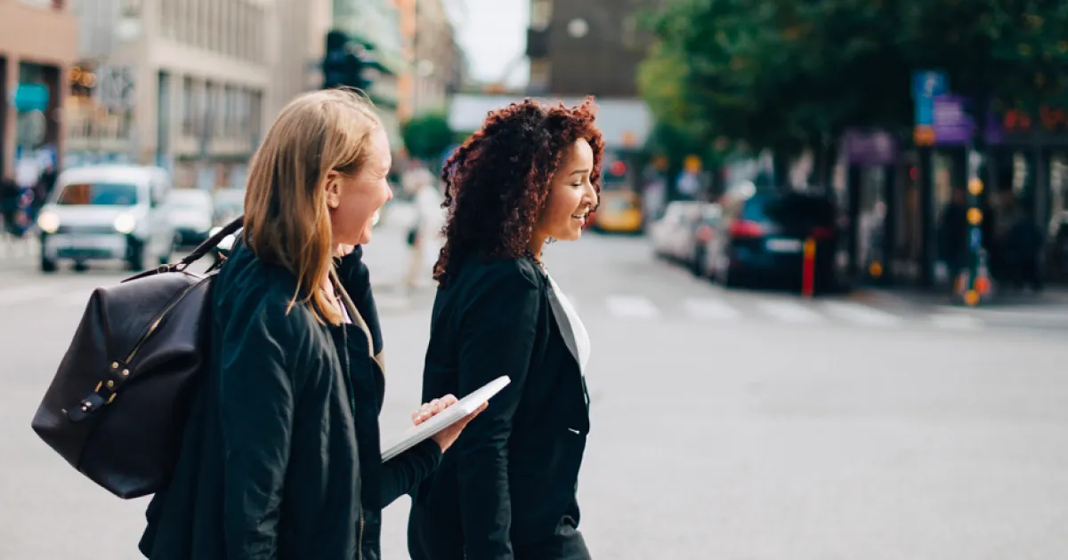 Två kvinnor promenerar på en soldränkt stadsgata. 