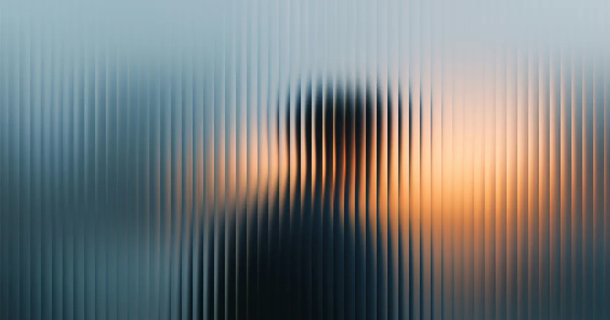 Silhouette of a person seen through vertically ribbed frosted glass with a sunset background.